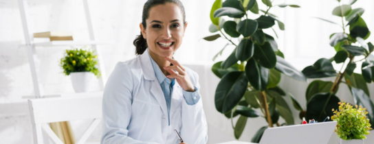 happy-nutritionist-in-white-coat-near-vegetables-and-laptop